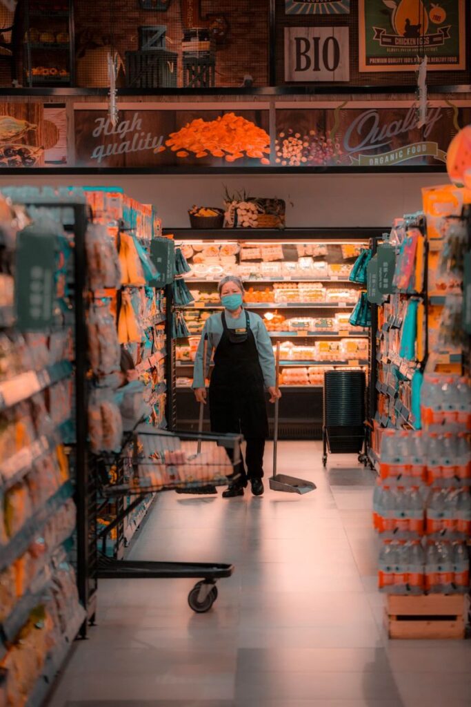 Elderly worker in mask cleans aisle of grocery store.