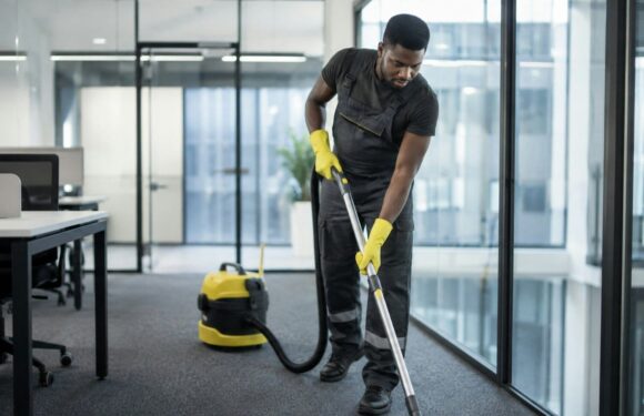 A man in yellow gloves vacuums a carpeted office floor.